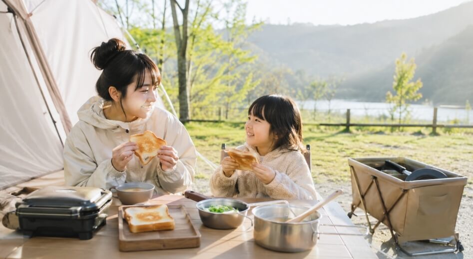 朝のキャンプ場で親子がホットサンドとスープの簡単な朝ごはんを楽しんでいる様子