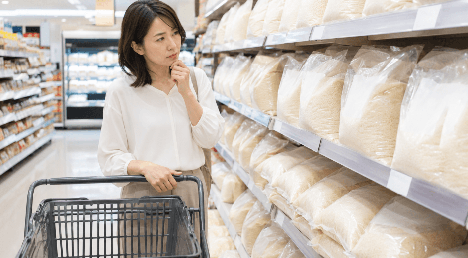 スーパーの米売り場で女性が米袋を見比べながら、安い日や価格を確認している様子