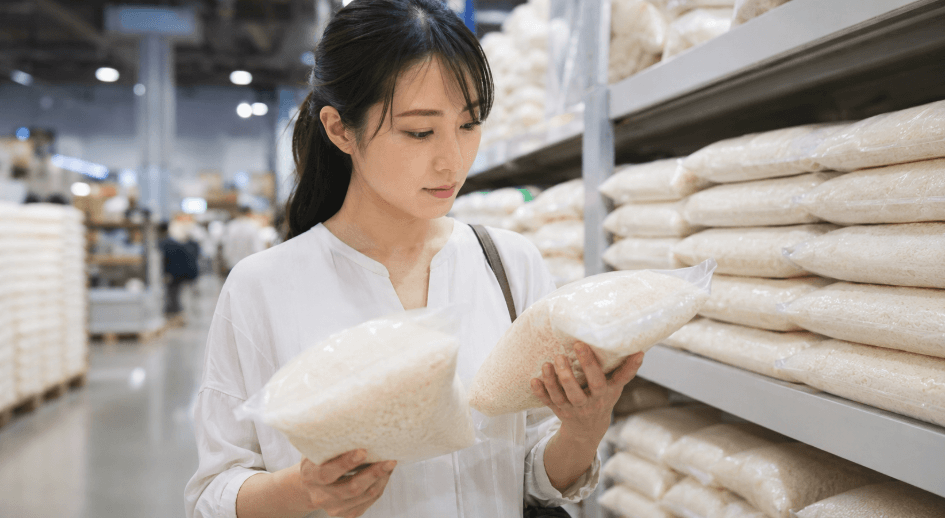 スーパーや量販店の米売り場で、さんさんまる米の価格や袋の状態を比較して選ぶ日本人女性